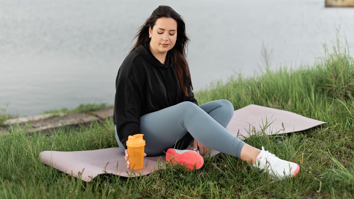 Woman enjoying a peaceful moment by the lake with a yoga mat and water bottle, promoting wellness.